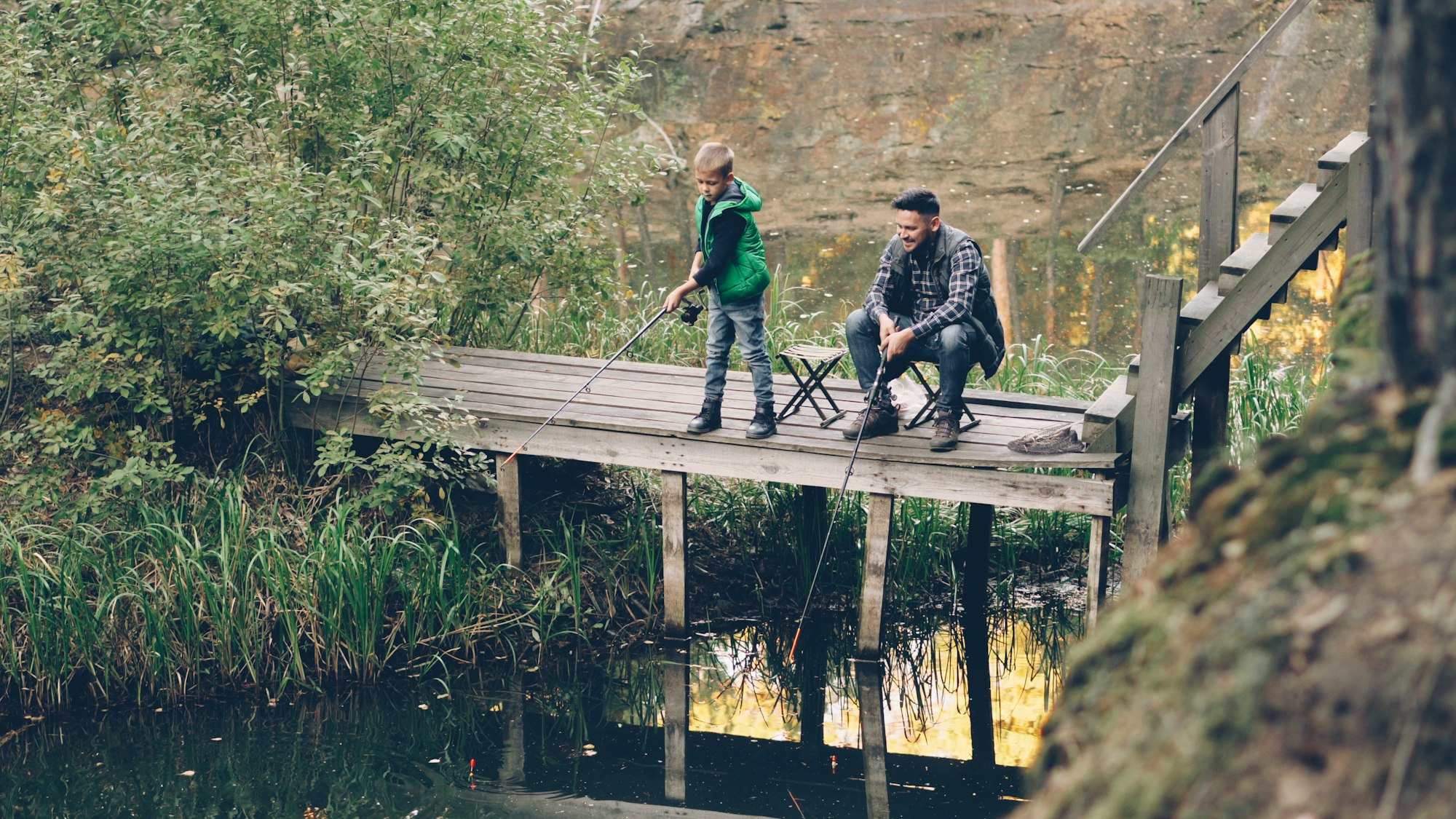 A kid casting a fishing line off a wooden dock while his dad coaches from beside him